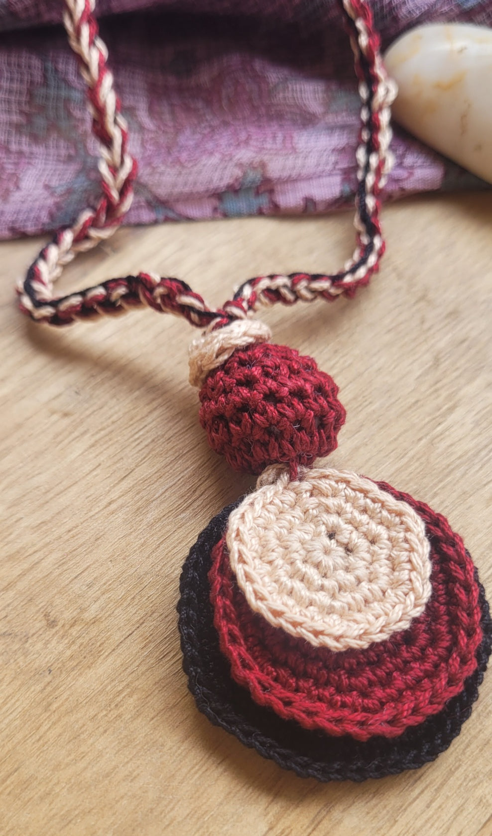 A handmade crocheted necklace with a layered pendant in red, cream, and black, resting on a light wood surface.