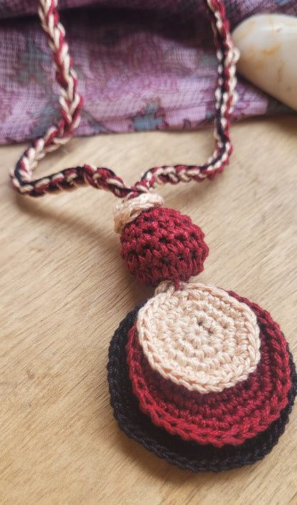 A handmade crocheted necklace with a layered pendant in red, cream, and black, resting on a light wood surface.