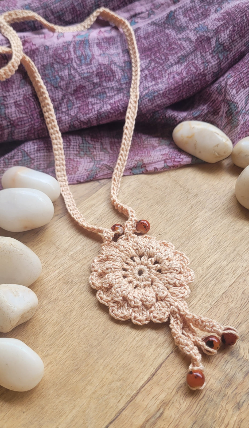 A bohemian-style crocheted necklace with a circular floral pendant and dangling tassels, made from light beige yarn with small brown beads.