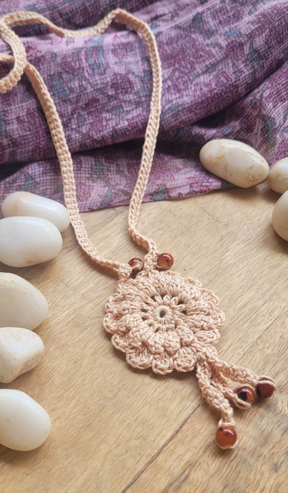 A bohemian-style crocheted necklace with a circular floral pendant and dangling tassels, made from light beige yarn with small brown beads.