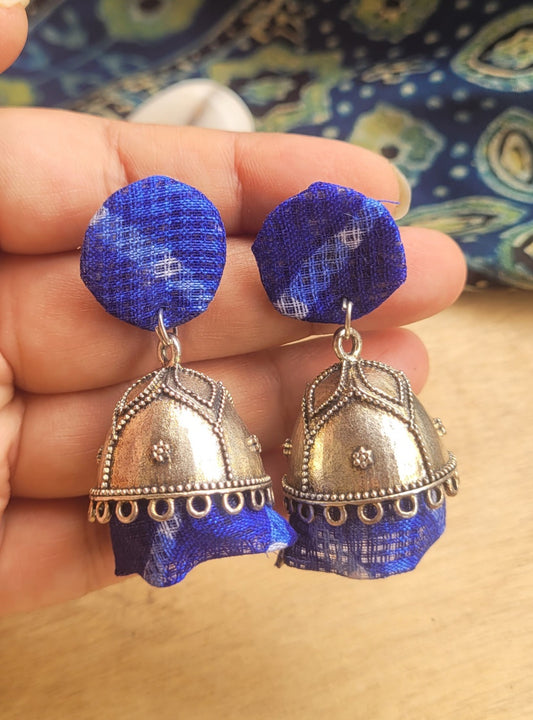 A pair of handmade Indian-style jhumka earrings. The earrings feature silver-toned metal jhumki decorated with silver beadwork, hanging from circular blue fabric studs with a subtle white pattern.