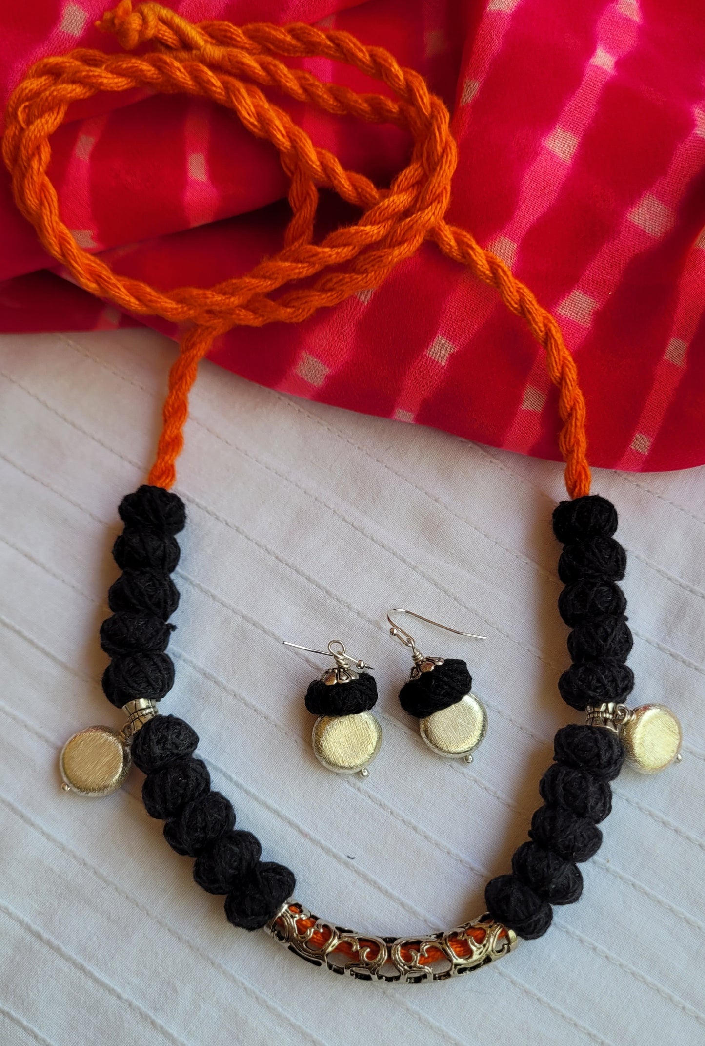 Black beaded necklace with gold accents and orange stones, paired with matching earrings, on a white surface with a red checkered fabric background.