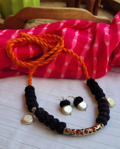 Black braided necklace with orange cord and matching earrings on a white surface with pink fabric in the background.